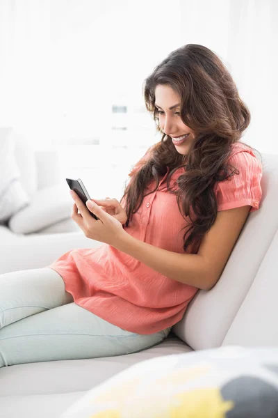 Happy brunette sitting on her couch using her smartphone — Stock Photo, Image