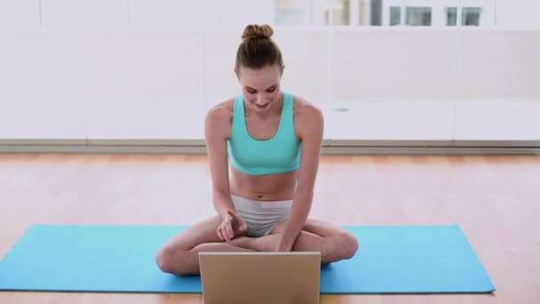 Подходящая модель сидит на коврике для упражнений, используя свой ноутбук — стоковое видео