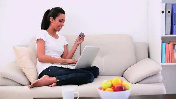 Mujer atractiva sentada en el sofá usando su computadora portátil y haciendo compras en línea — Vídeos de Stock