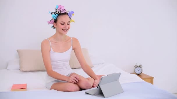 Jovem modelo em rolos de cabelo usando seu tablet digital para vídeo chat em sua cama — Vídeo de Stock