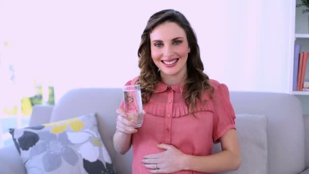 Ganska gravid modell sitter på hennes soffa dricka glas vatten — Stockvideo