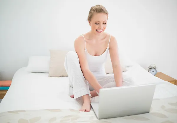 Естественная счастливая блондинка смотрит на ноутбук, сидя на кровати — стоковое фото
