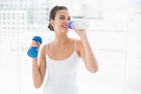 Frau in weißer Sportbekleidung trinkt Wasser, während sie eine Hantel hebt — Stockfoto