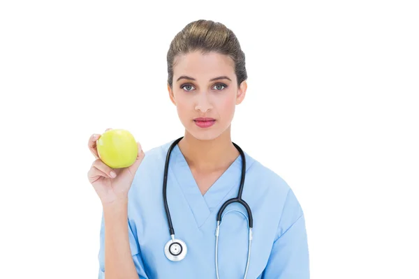 Serious nurse in blue scrubs holding a green apple — Stock Photo, Image