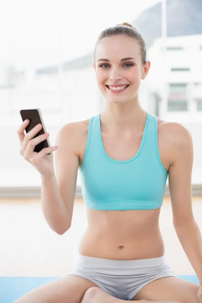Sporty smiling woman holding phone while sitting on exercising mat — Stock Photo, Image