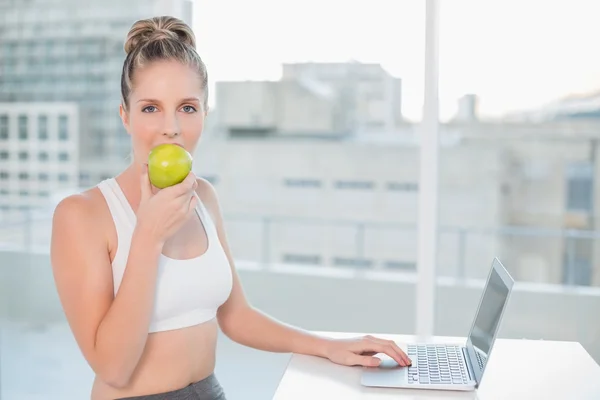 Loira desportiva comendo maçã ao usar laptop — Fotografia de Stock