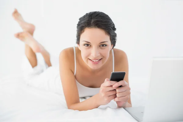 Entzückte lässige Brünette im weißen Pyjama mit ihrem Handy auf ihrem Bett liegend — Stockfoto