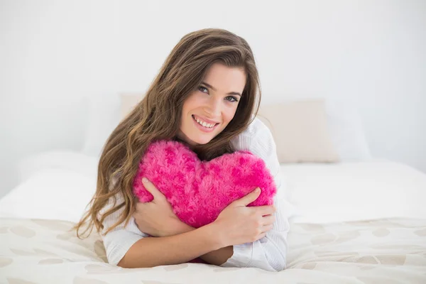 Mulher agradável em pijama branco abraçando um travesseiro em forma de coração — Fotografia de Stock