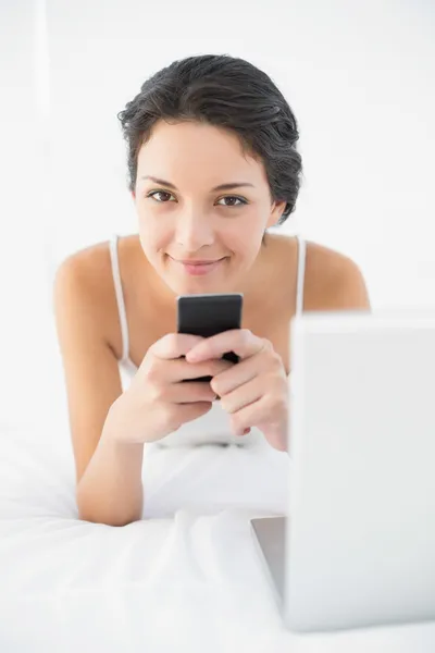Ontspannen casual brunette in witte pyjama's met behulp van haar mobiele telefoon — Stockfoto