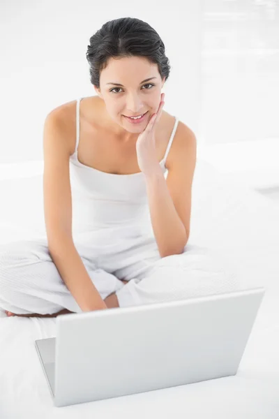 Fröhliche lässige Brünette im weißen Pyjama sitzt auf ihrem Bett und benutzt einen Laptop — Stockfoto