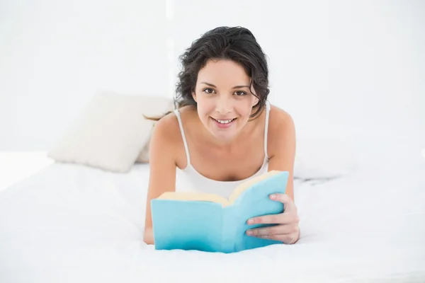 Lächelnde Brünette im weißen Pyjama entspannt sich auf einem Bett und liest ein Buch — Stockfoto