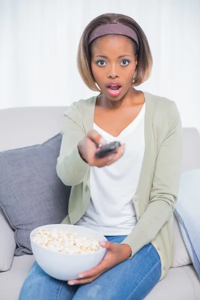 Mulher chocada sentado no sofá mudando canal de tv enquanto segurando pipocas — Fotografia de Stock