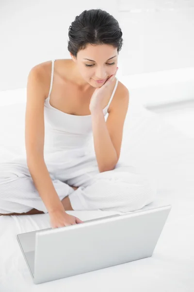 Concentrado casual morena em pijama branco sentado em sua cama e usando um laptop — Fotografia de Stock