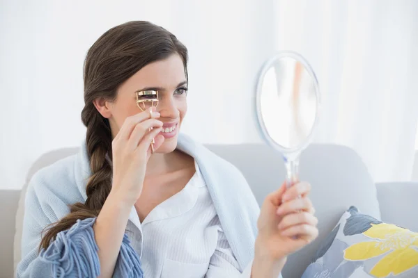 Concentrated woman in white pajamas using an eyelash curler — Stock Photo, Image