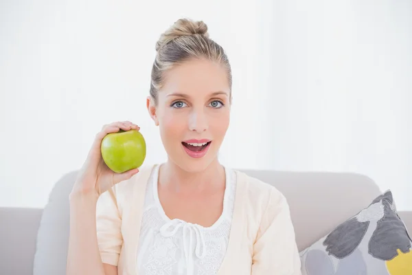 Surprised gorgeous model holding green apple sitting on sofa — Stock Photo, Image