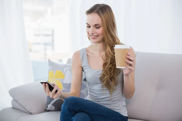 Mujer joven sentada en el sofá sosteniendo mensajes de texto taza desechable en el teléfono — Foto de Stock