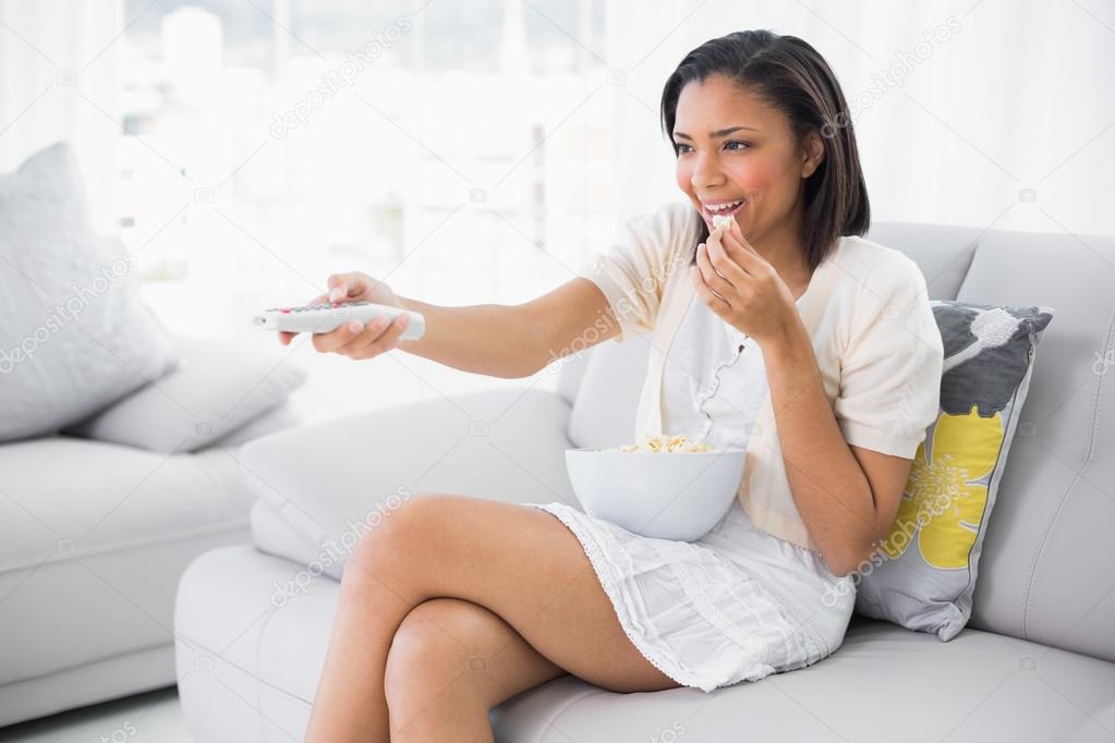Pleased young woman in white clothes eating popcorn while watching tv