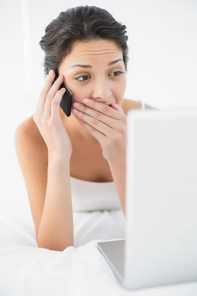 Verrast casual brunette in witte pyjama's maken van een telefoongesprek — Stockfoto