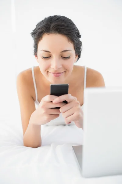 Joyful casual morena de pijama branco usando seu telefone celular deitado em sua cama — Fotografia de Stock