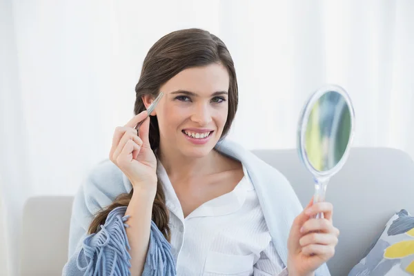 Smiling woman in white pajamas plucking her eyebrows — Stock Photo, Image