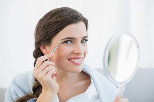 Happy woman in white pajamas plucking her eyebrows — Stock Photo, Image