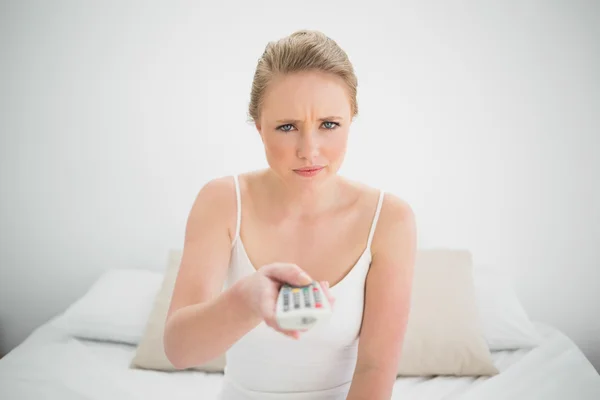 Natural frowning blonde holding remote while sitting on bed — Stock Photo, Image