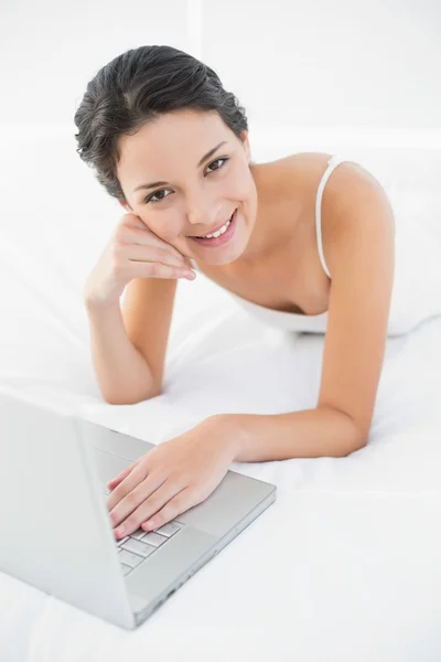 Smiling casual brunette in white pajamas using a laptop — Stock Photo, Image