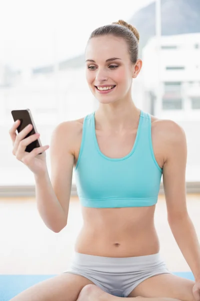 Deportiva mujer alegre sosteniendo el teléfono mientras está sentado en la esterilla de ejercicio — Foto de Stock