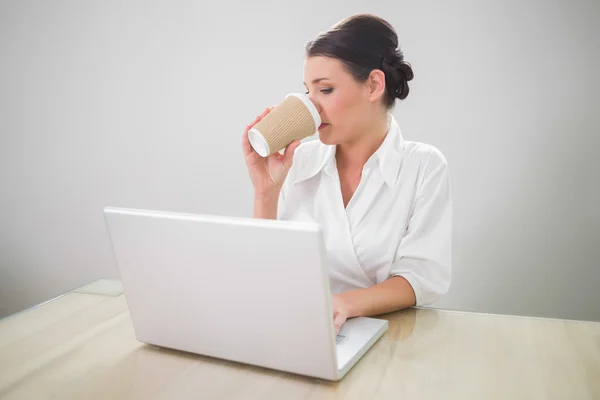 Gorgeous businesswoman drinking coffee while using laptop — Stock Photo, Image