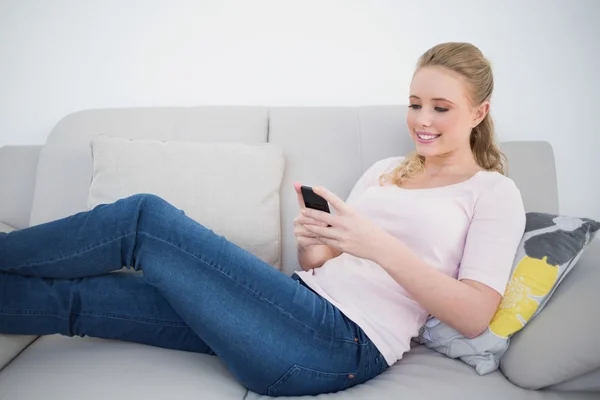 Casual smiling blonde using smartphone and lying on couch — Stock Photo, Image