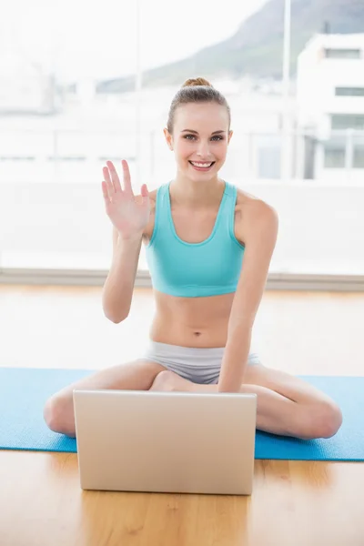 Sportig leende kvinna sitter med benen i kors framför laptop vinka — Stockfoto