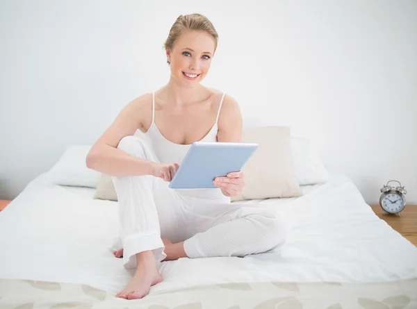 Natural sonriente rubia sosteniendo la tableta mientras está sentado en la cama —  Fotos de Stock