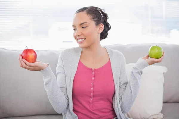 Веселая женщина сидит на диване, держа зеленое и красное яблоко, глядя на красное. — стоковое фото