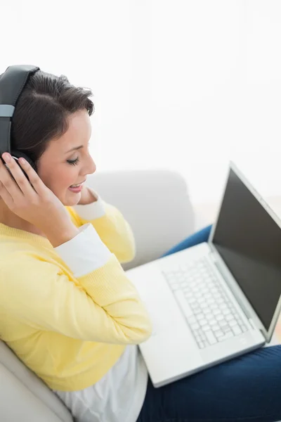 Glad casual brunett i gula cardigan lyssna på musik medan du använder en bärbar dator — Stockfoto