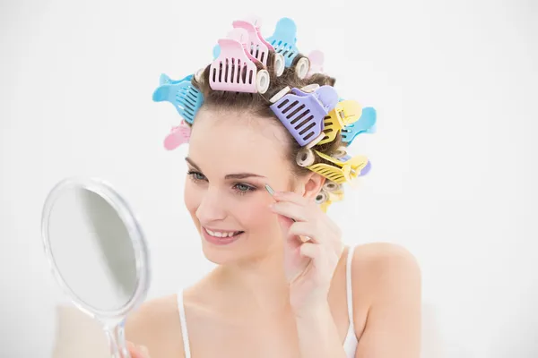Cute woman in hair curlers plucking her eyebrows — Stock Photo, Image