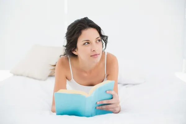 Pensativa morena casual en pijama blanco relajándose en una cama leyendo un libro — Foto de Stock