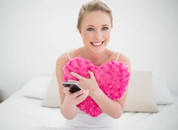 Natural cheerful blonde holding heart pillow and smartphone — Stock Photo, Image