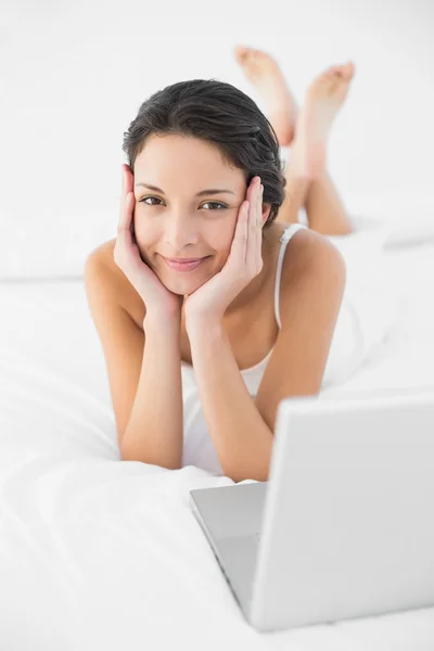 Happy casual brunette in white pajamas lying on her bed and looking at camera — Stock Photo, Image