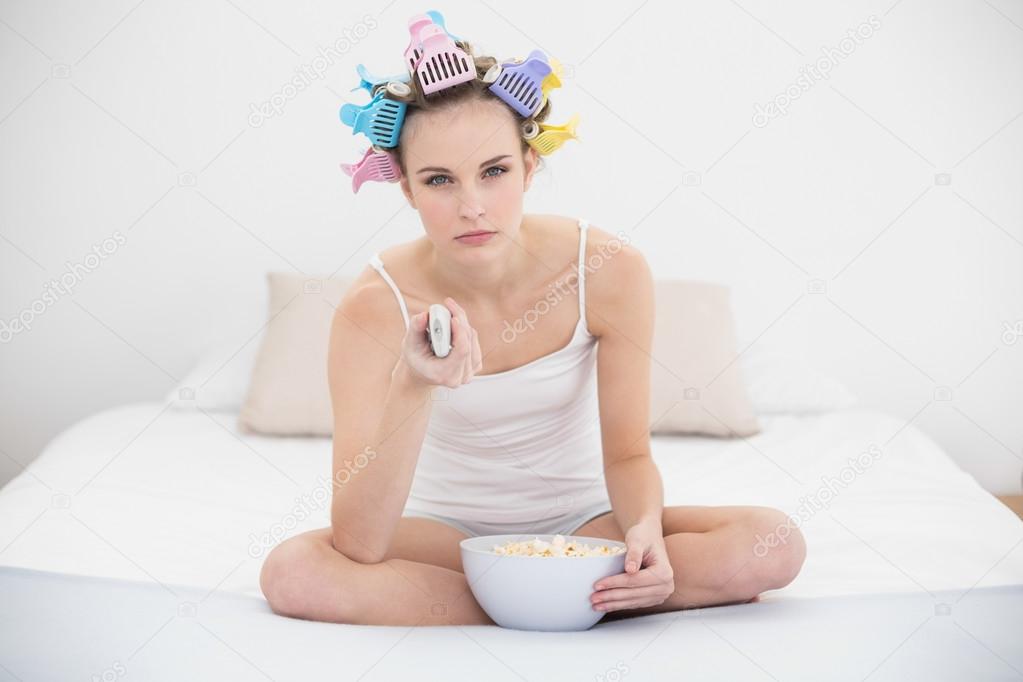 Bored woman in hair curlers watching tv while eating popcorn