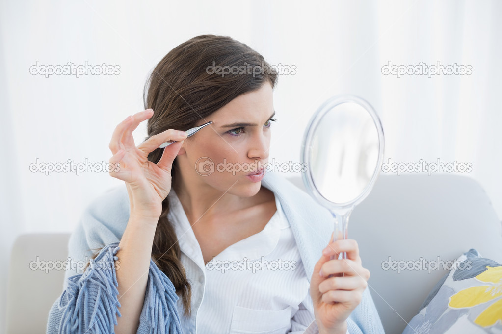 Focused woman in white pajamas plucking her eyebrows