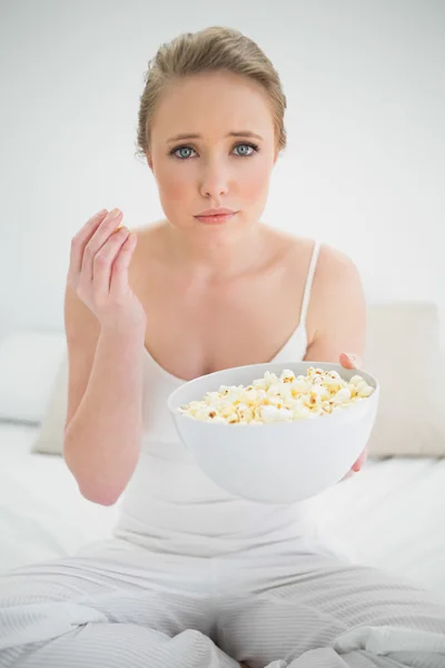 Natural frowning blonde holding bowl of popcorn on bed — Stock Photo, Image