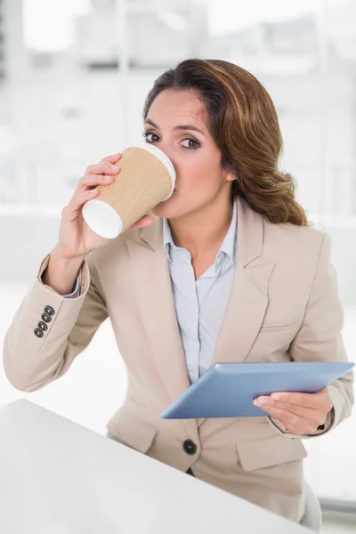 Mujer de negocios sonriente usando tableta digital en su escritorio bebiendo de la taza desechable —  Fotos de Stock