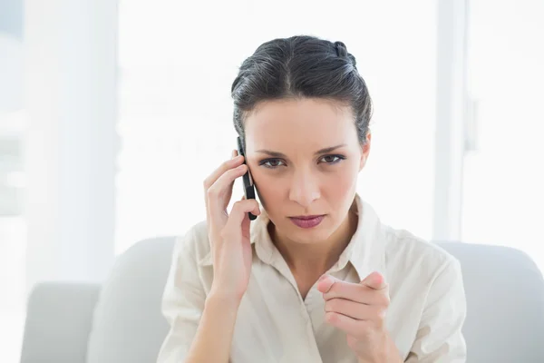 Irritada mujer de negocios morena elegante haciendo una llamada telefónica y apuntando cámara con su dedo —  Fotos de Stock