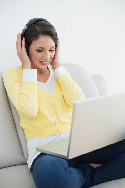 Relaxed casual brunette in yellow cardigan listening to music while using a laptop — Stock Photo, Image