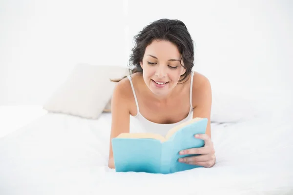 Encantadora morena casual de pijama branco deitada em sua cama lendo um livro — Fotografia de Stock