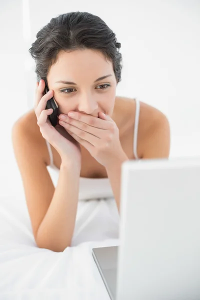 Happy casual brunette in white pajamas making a phone call and looking at her laptop — Stock Photo, Image