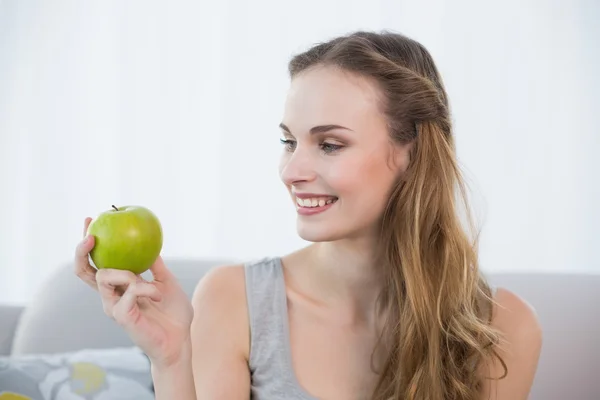 Mujer joven sonriente sentada en un sofá sosteniendo manzana verde —  Fotos de Stock