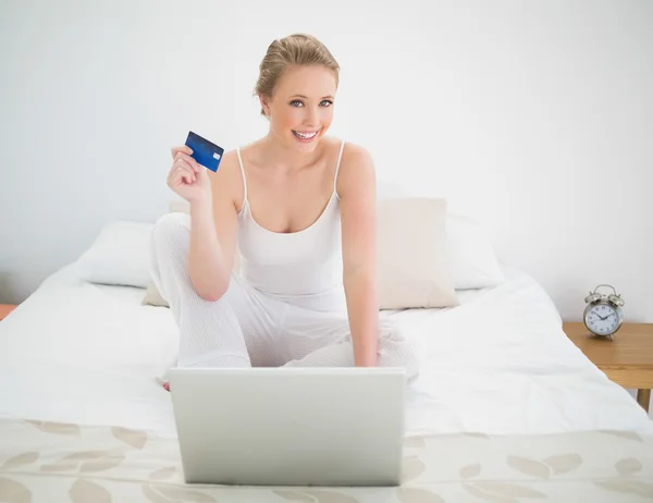 Natural cheerful blonde holding credit card and sitting in front of laptop — Stock Photo, Image