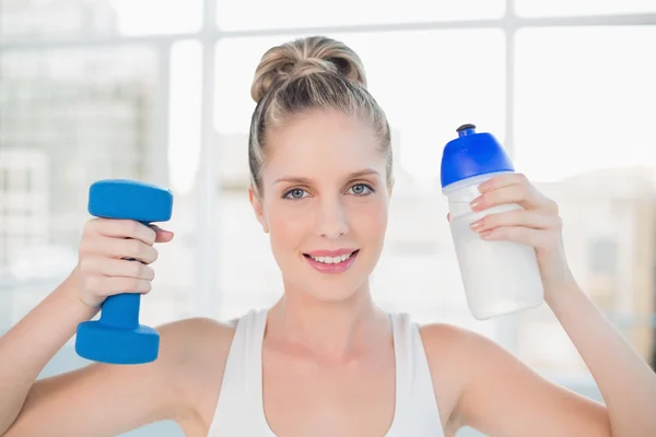 Smiling sporty blonde holding flask and dumbbell — Stock Photo, Image