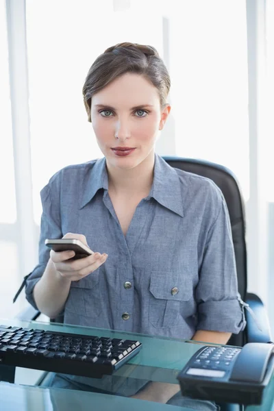 Empresaria sosteniendo su smartphone sentada en su escritorio —  Fotos de Stock